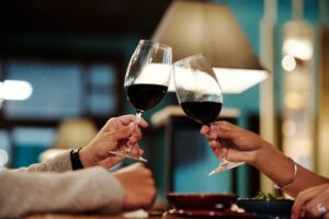 Two people enjoying a romantic dinner, clinking red wine glasses in a cozy restaurant setting.